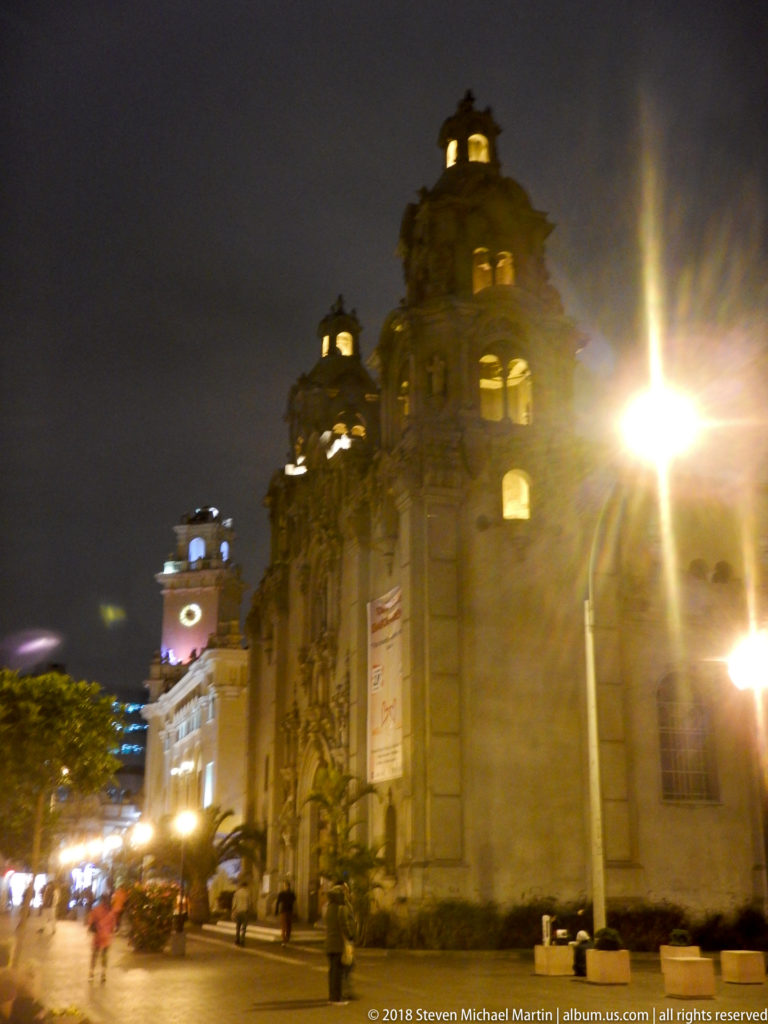 Ornate illuminated civic buildings and a clock tower along a Miraflores street at night, with pedestrians and bright streetlamps.