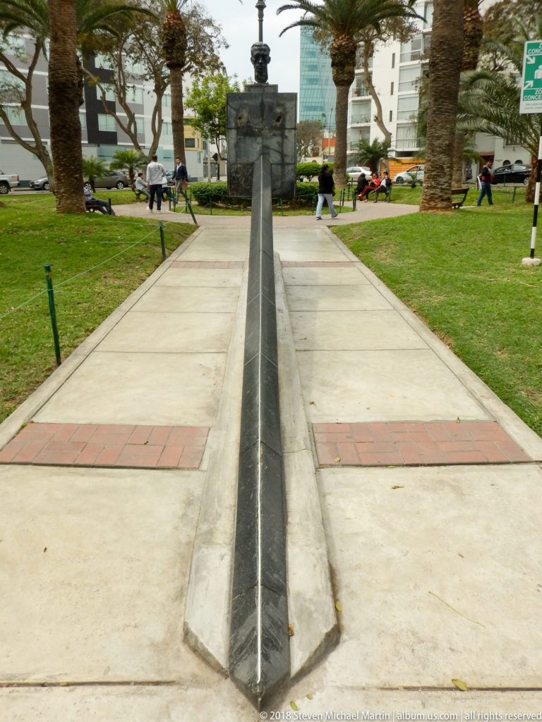 Long narrow dark-stone channel aligned with a small upright monument in a palm-lined urban park.