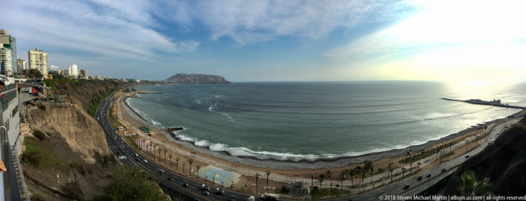 High view over Miraflores cliffs, the Costa Verde roadway, narrow beach, Pacific surf, and distant headland.