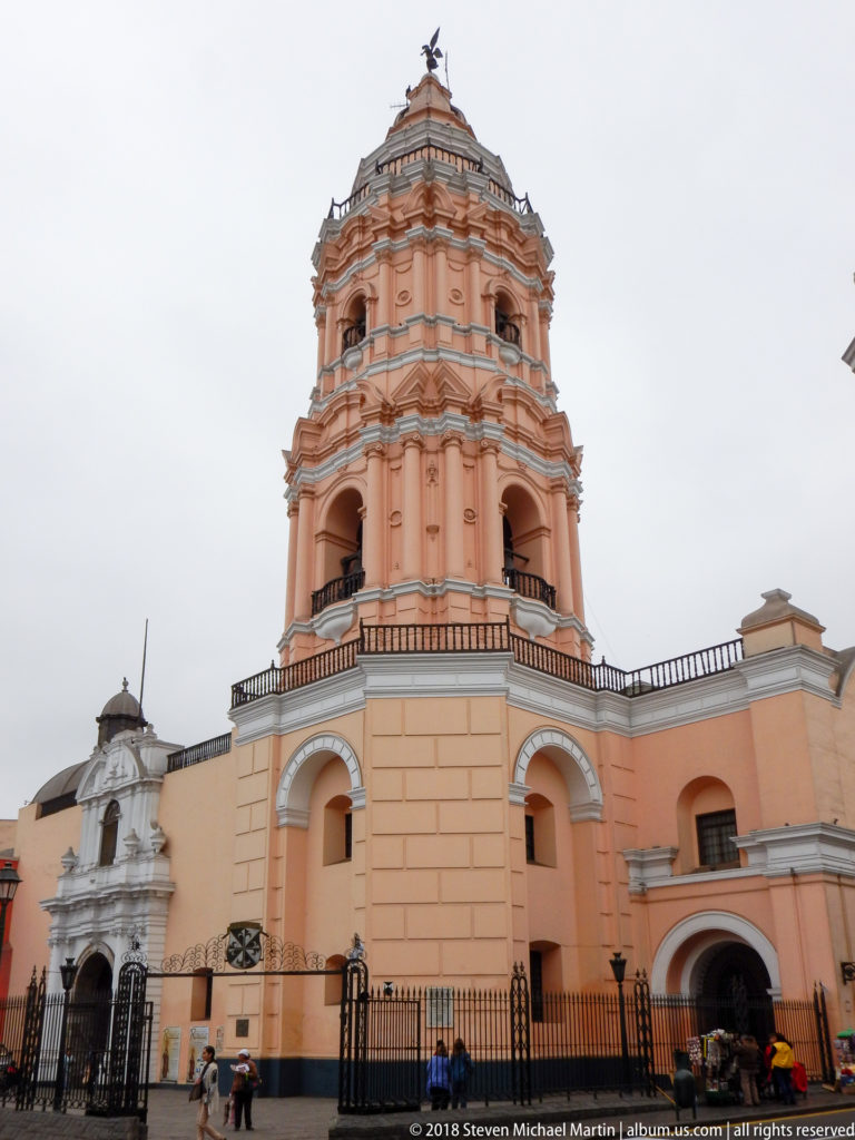 Street-level view of Santo Domingo’s tall peach bell tower with arched belfry openings, layered moldings, and adjoining church walls.