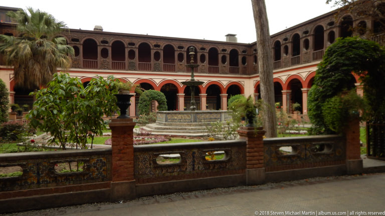 Main cloister garden with tiled low wall, central fountain, trees, and two-story red-and-cream arcaded galleries.