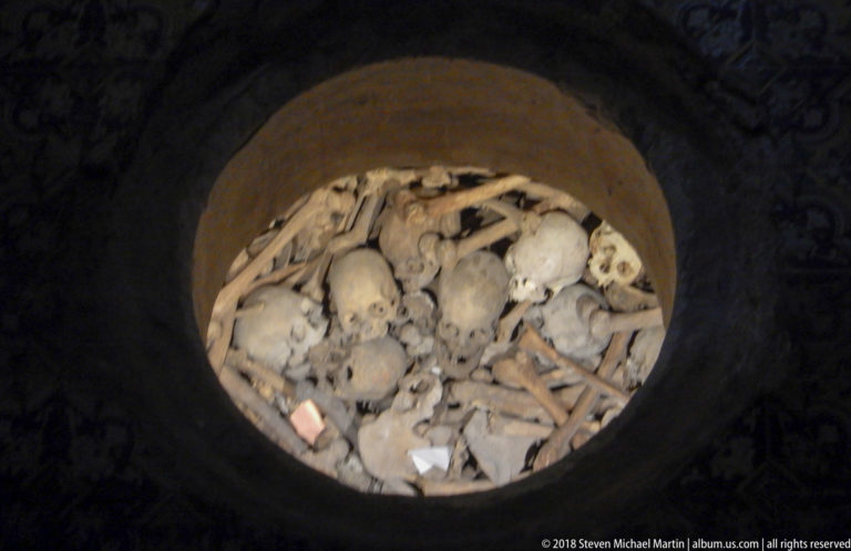 Circular crypt opening filled with commingled human skeletal remains, including several skulls and long bones.
