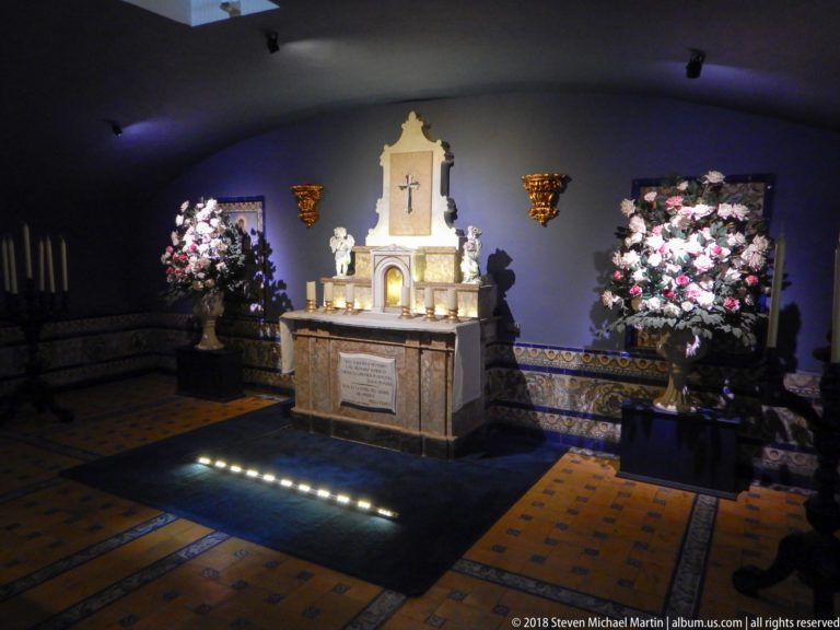 Small illuminated altar in a low vaulted crypt, flanked by flower arrangements with a memorial slab at the front.