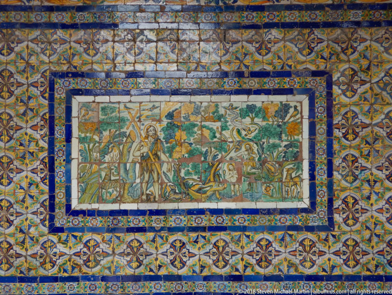 Figurative ceramic tile mural with a haloed religious figure, plants, animals, and people surrounded by blue-and-yellow patterned borders.