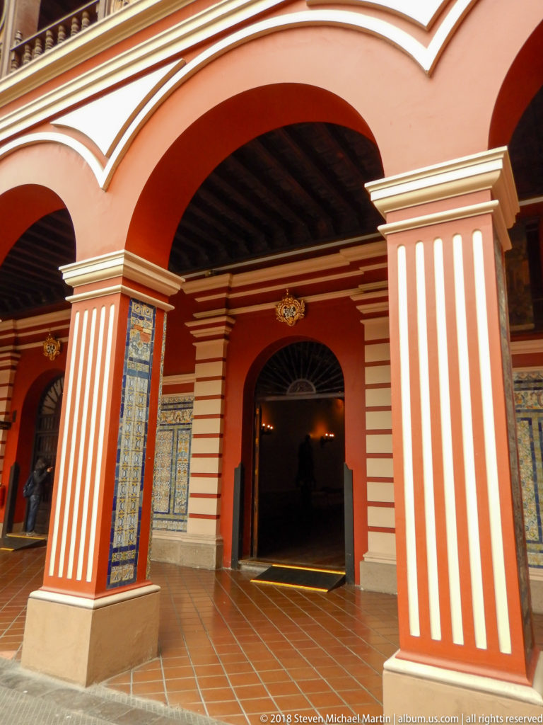 Red-and-white cloister arcade with broad arches, striped piers, blue-and-white tile panels, terracotta floor, and dark timber ceiling.