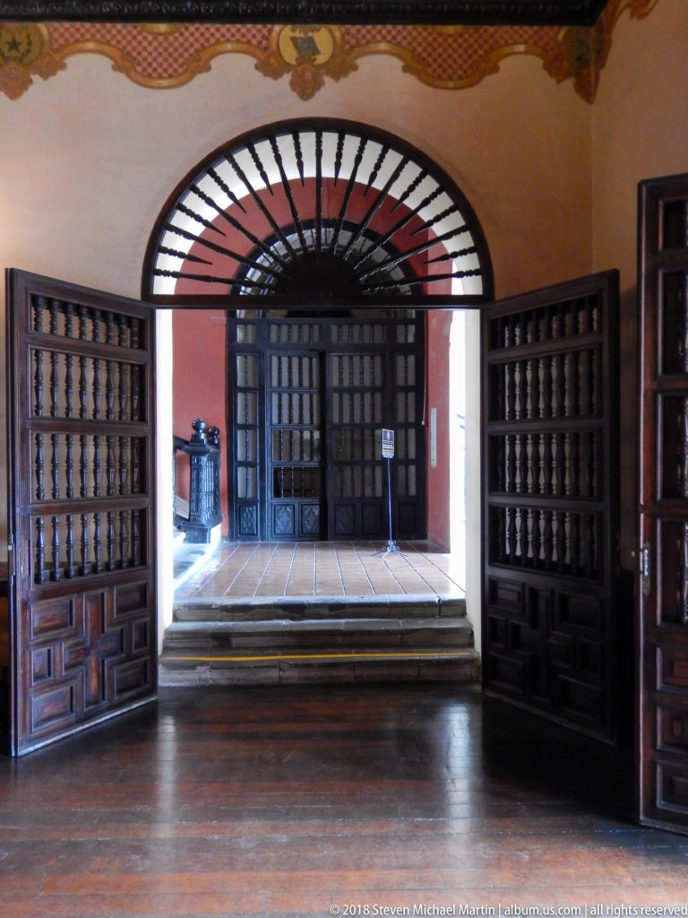 Open heavy wooden doors beneath an arched transom lead from a dim interior across a raised threshold into a brighter passage.