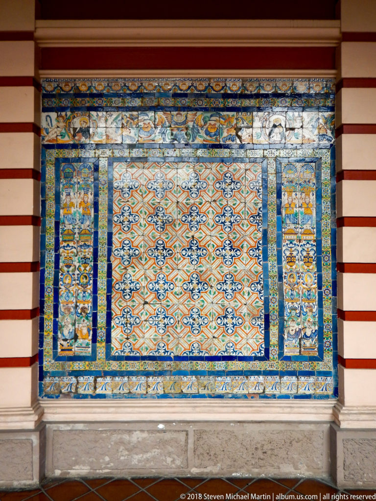 Large blue, orange, yellow, green, and white glazed-tile panel set into a red-and-white cloister wall.