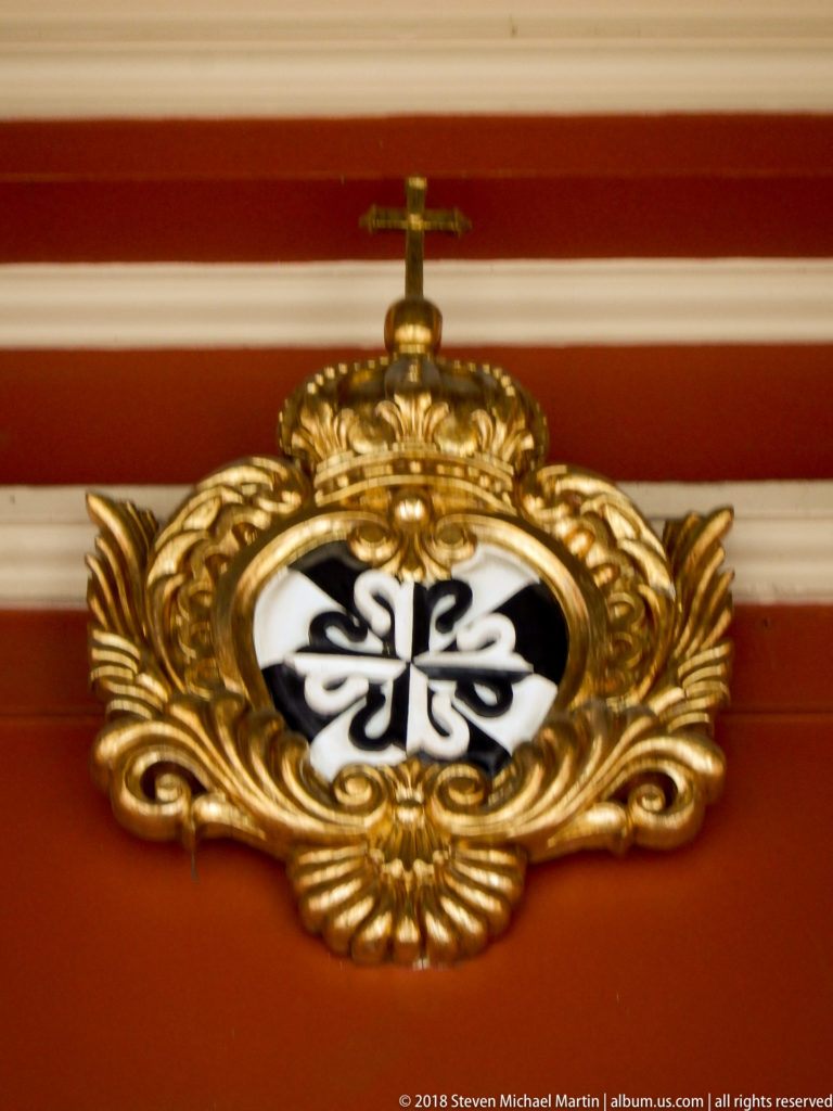 Black-and-white Dominican cross set within an elaborate gilded scrolling cartouche on a red wall.
