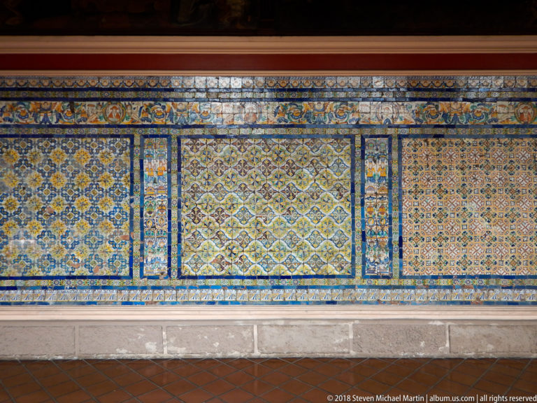 Large cloister wall covered by blue, yellow, green, and white patterned glazed tiles arranged in framed rectangular panels.