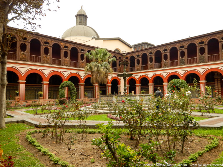 Wide view across Santo Domingo’s cloister garden toward red arcades, central fountain, palm trees, and a pale dome.