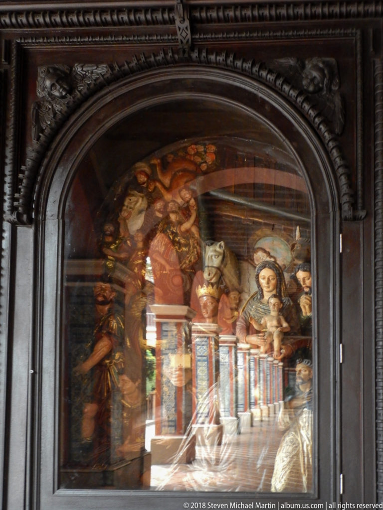 Large framed religious painting behind reflective glass, with several sacred figures partly obscured by reflections of the cloister.