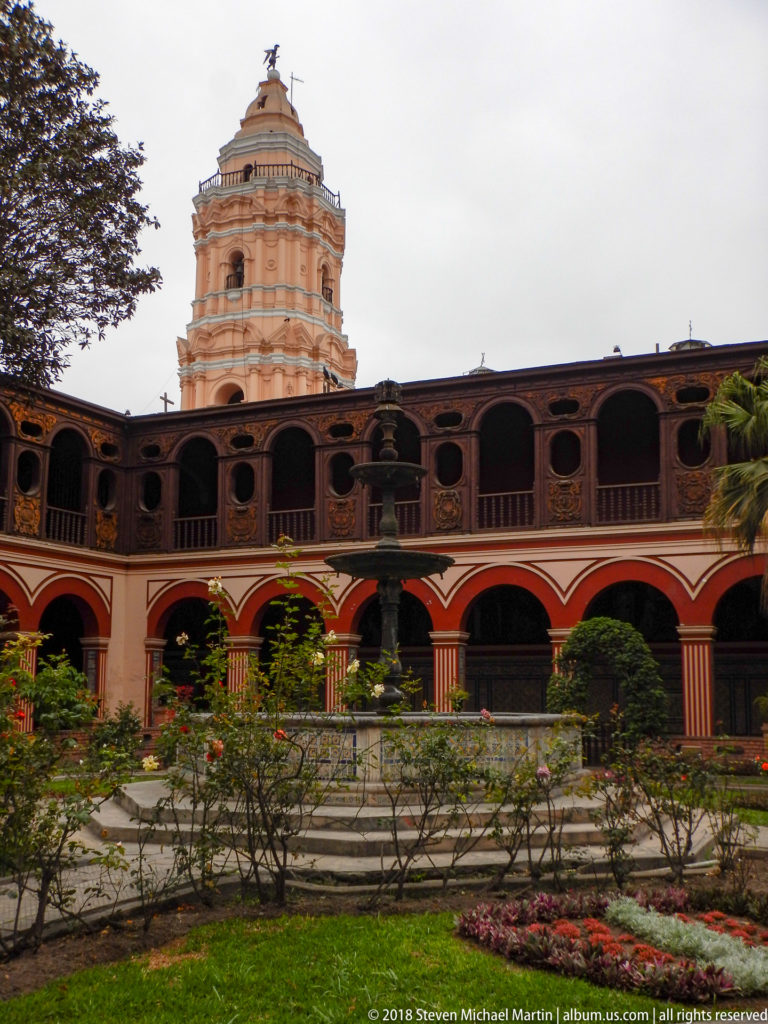 Centered cloister garden view with roses, dark fountain, two-level arcades, and Santo Domingo bell tower rising behind.