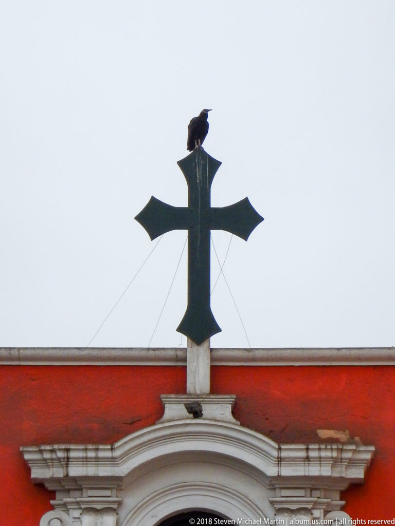 Dark metal cross above a white curved pediment on a red wall, with a bird perched on its upper arm.