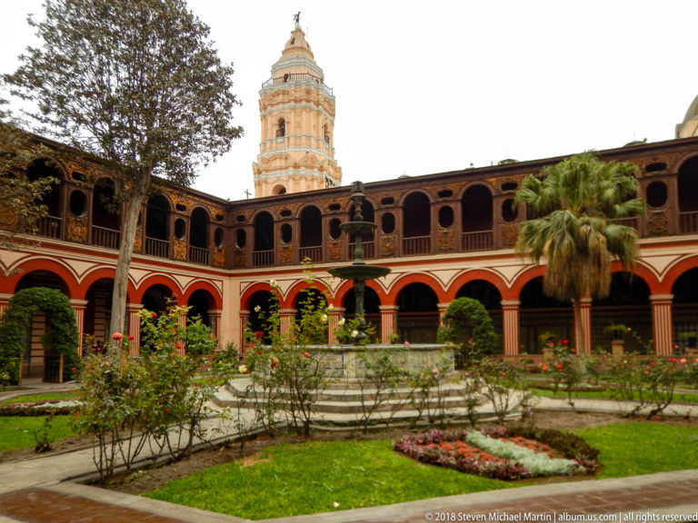 Main cloister garden with flowers, fountain, two-story red-and-cream arcades, palms, and Santo Domingo bell tower behind.