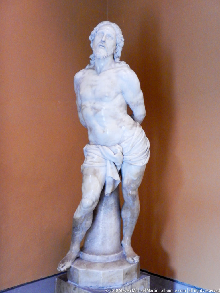 Pale stone statue of a semi-nude male figure with draped cloth on a cylindrical support against an ochre wall.