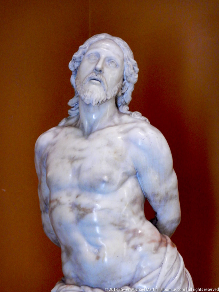 Close view of a white marble bound male figure with bare torso, uplifted head, and arms restrained behind the back.