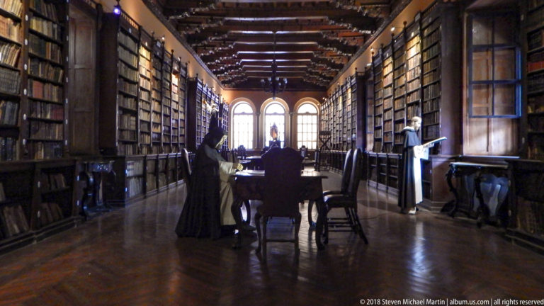 Long view through Santo Domingo library with floor-to-ceiling bookcases, timber ceiling, reading tables, and three arched windows.