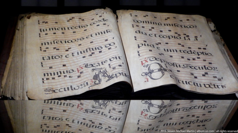 Close view of an open choir book with black square notation, red staff lines, liturgical text, and reflection in glass.