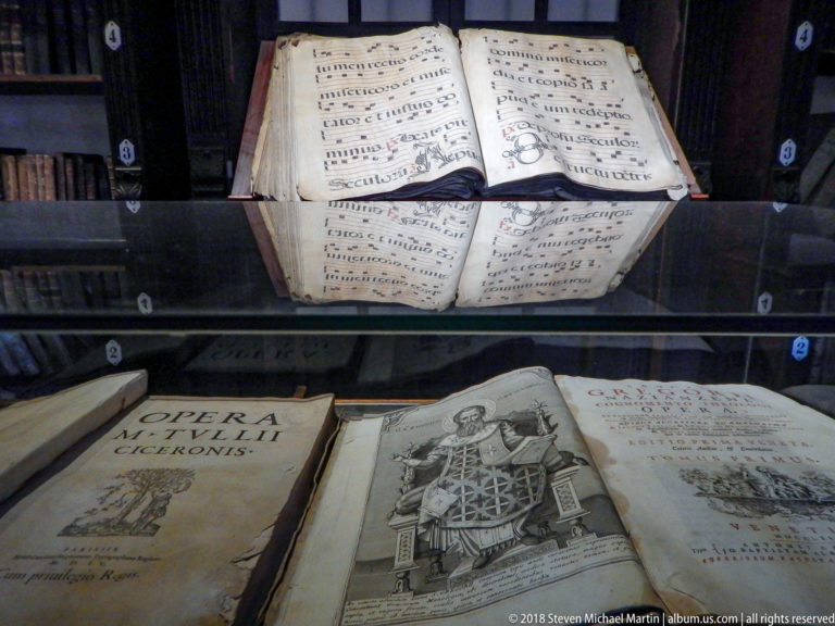 Glass case displaying an open choir book above historic printed volumes with a title page and engraved illustration.