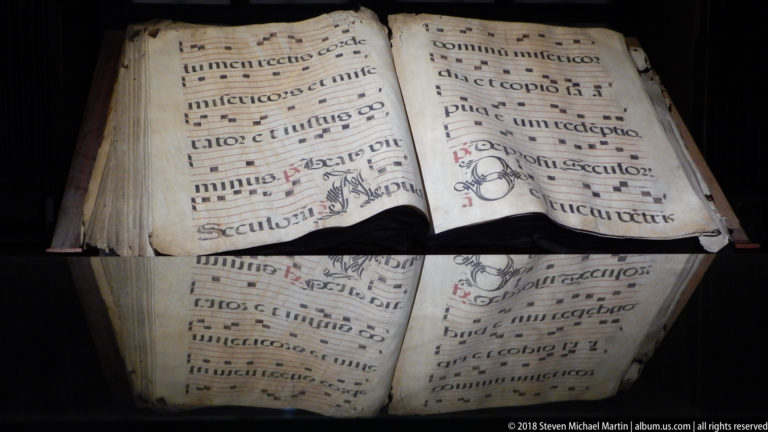 Open choir book with large musical notation and liturgical text, strongly reflected in the protective display glass below.