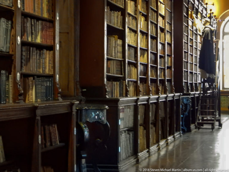 Historic library interior with tall dark wooden bookcases, bound volumes, reading furniture, and polished floor.