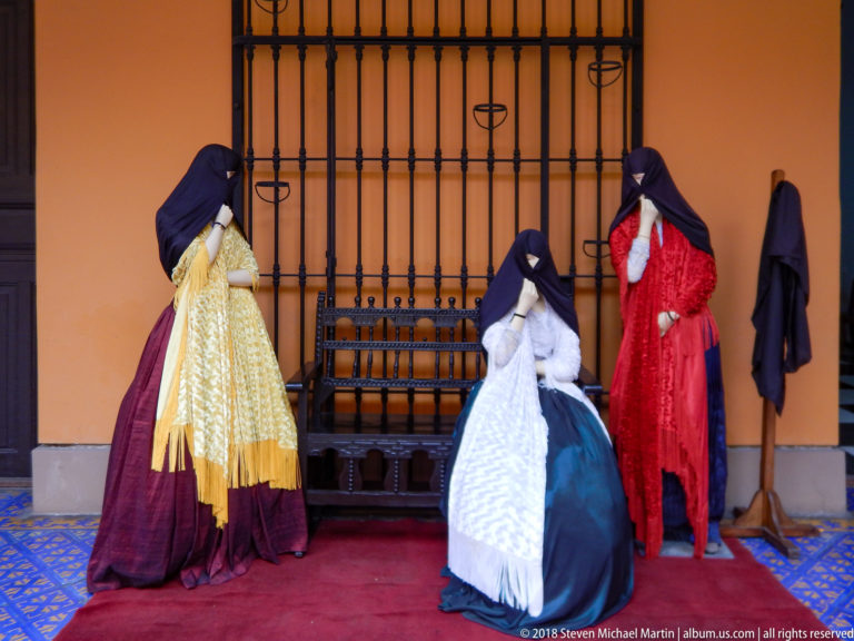 Three mannequins in colorful tapada costumes with dark face-covering mantles, displayed before an iron grille.