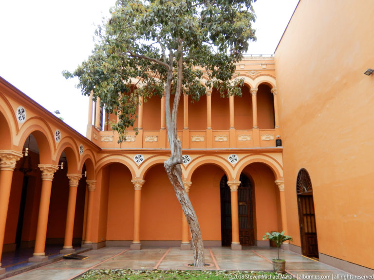 Two-story orange arcaded courtyard with a mature tree, paved floor, and simple doors and windows.