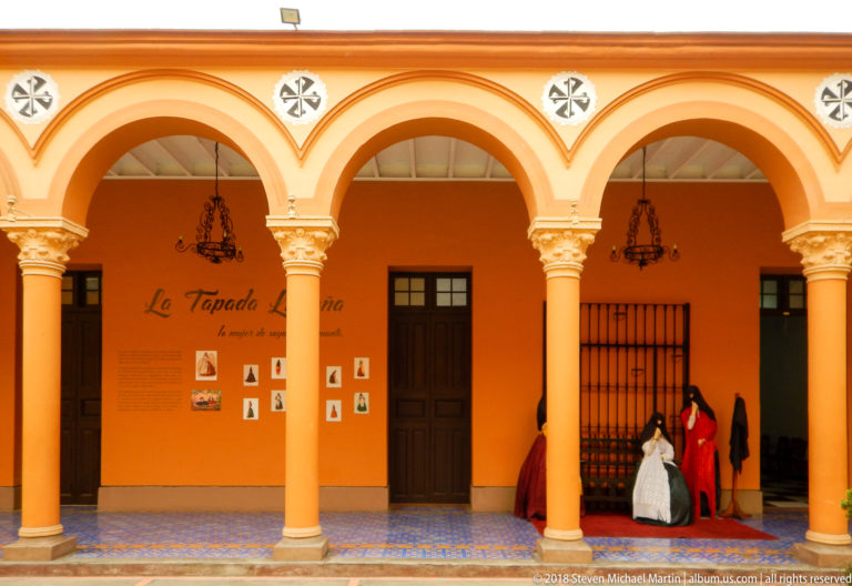 Orange cloister arcade containing the La Tapada Limeña panel and mannequins in historic covered dress.