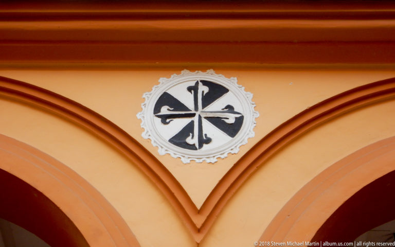 Black-and-white Dominican cross medallion centered in the spandrel above two orange cloister arches.