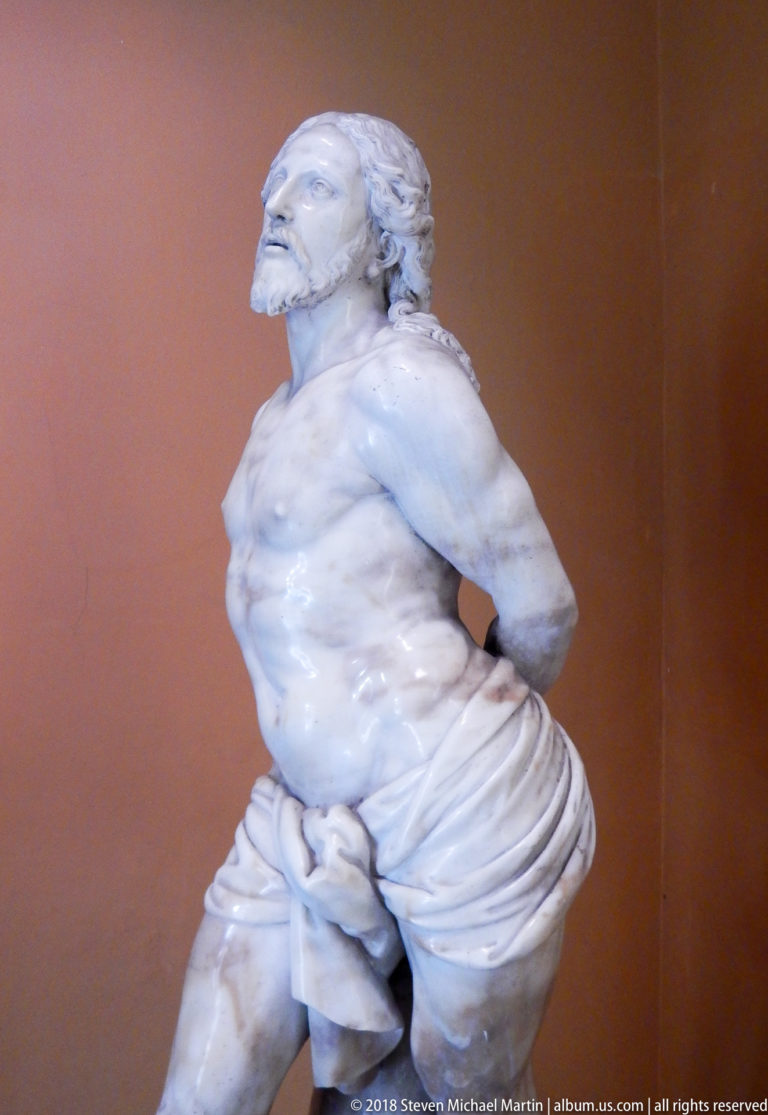 White marble sculpture of a bound, wounded man with bare torso and arms restrained behind his back.