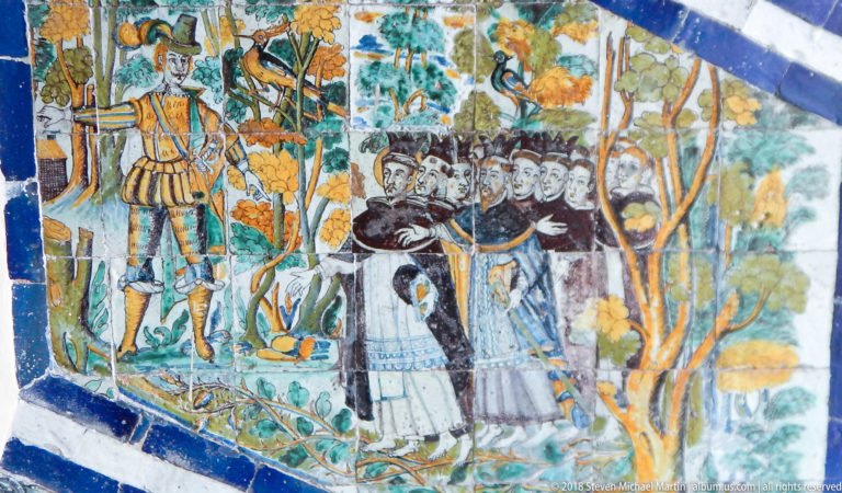 Painted ceramic tile panel showing friars and armed figures among trees, bordered in blue.