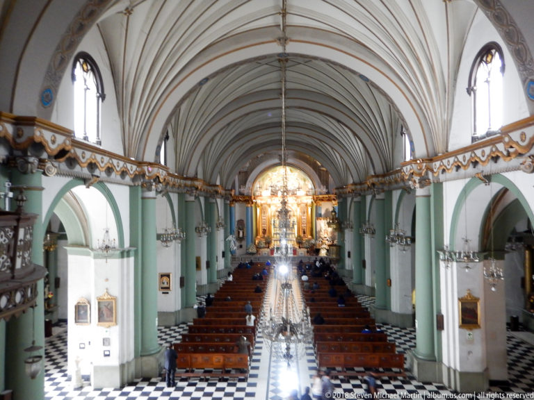 Elevated centered view down Santo Domingo’s nave with ribbed vault, green-white piers, pews, patterned floor, and bright high altar.