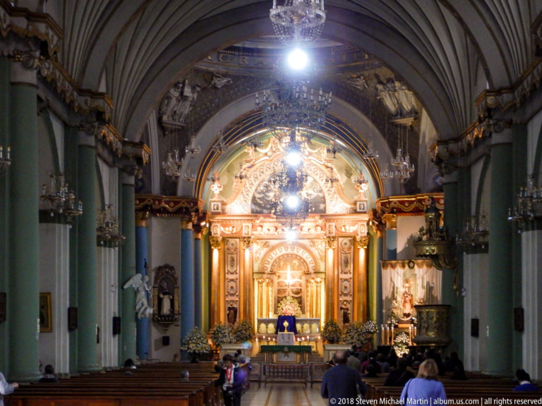 People seated in Santo Domingo’s nave facing the illuminated high altar beneath chandeliers and a ribbed vaulted ceiling.