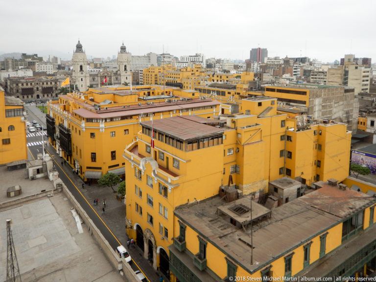 Elevated view across ochre colonial buildings and streets in central Lima, with church towers and modern skyline beyond.