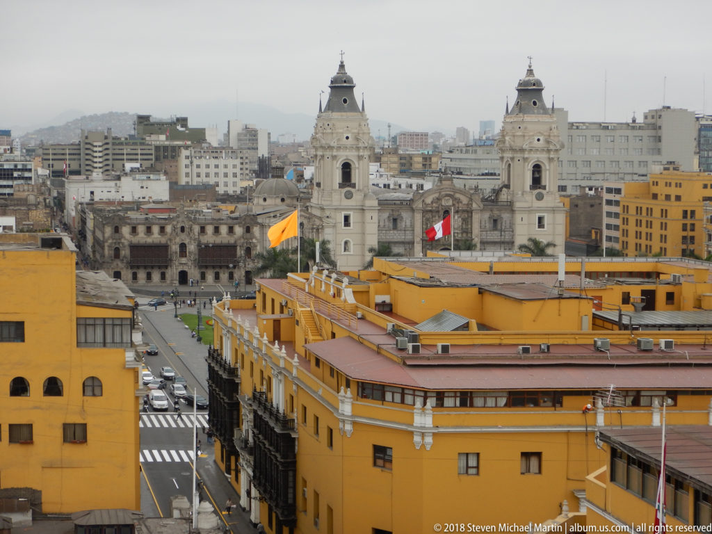 Two pale church towers rising over central Lima, with ochre buildings, flags, streets, and distant urban skyline.