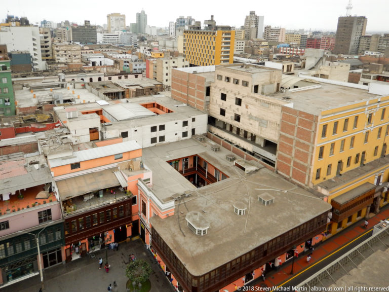 Elevated view over central Lima with colorful street façades, inner courtyards, flat roofs, and taller modern buildings beyond.