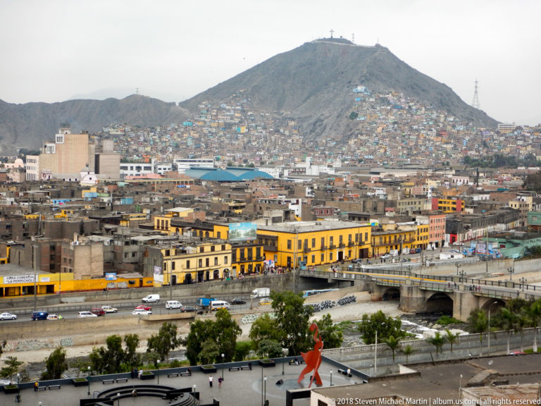 Elevated view from Santo Domingo over central Lima, with yellow historic façades, roadways, dense hillside housing, and a steep hill.