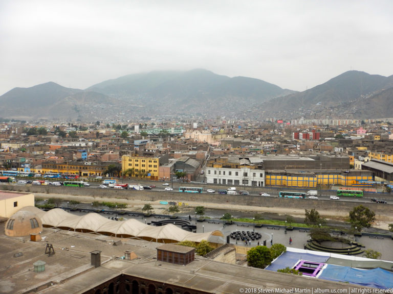 Elevated Lima panorama with dense low-rise buildings, roadway, foreground roofs, and mist-covered hills.
