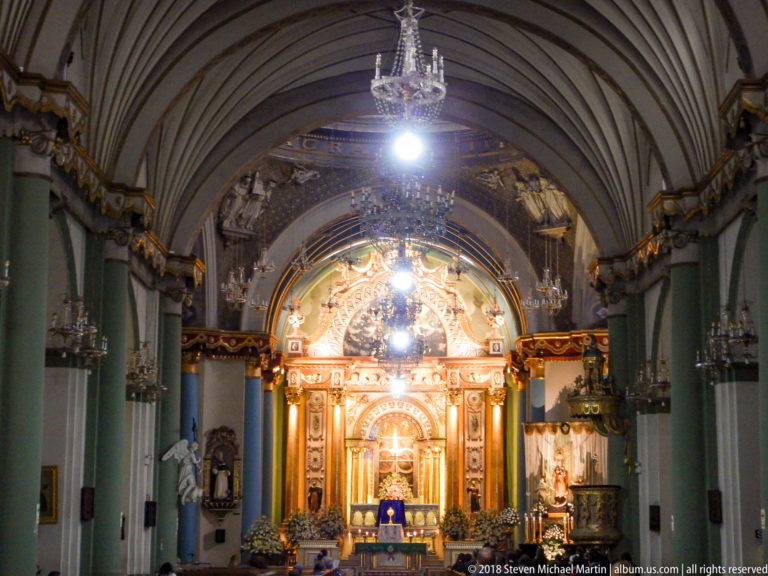 Axial view down Santo Domingo’s nave toward a brightly illuminated gilded high altar beneath a ribbed vault and chandeliers.