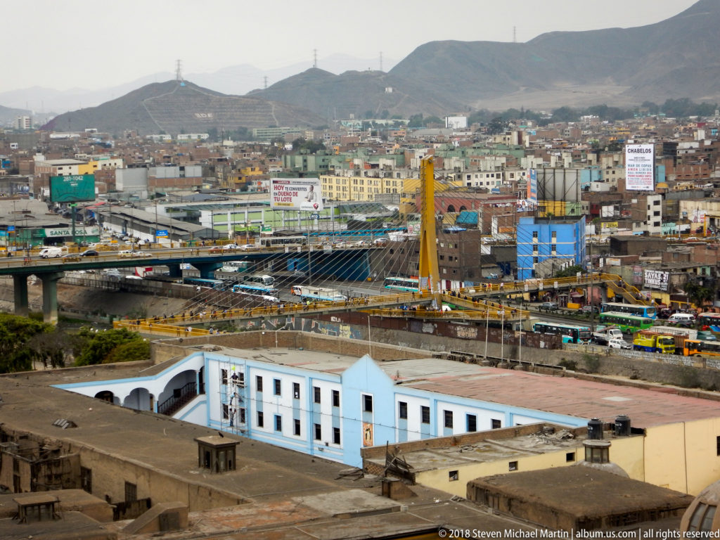 Tower view over Lima toward a yellow-pylon cable-stayed bridge, dense hillside neighborhoods, and a white-blue building.