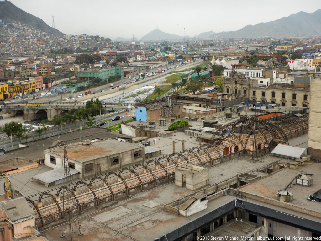 Elevated panorama over central Lima from Santo Domingo, with rooftops, bridge, roads, historic buildings, and distant hills.