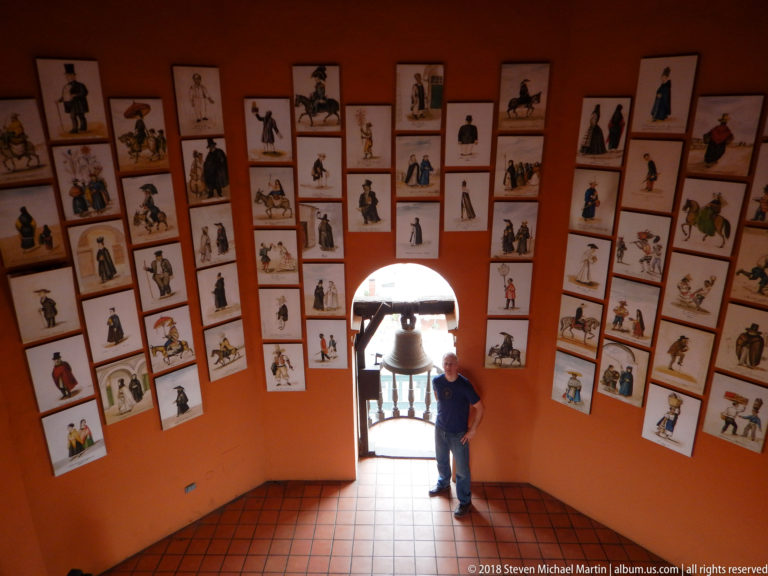 Visitor standing in an orange tower room covered with historical figure prints, with a bell visible through the central opening.