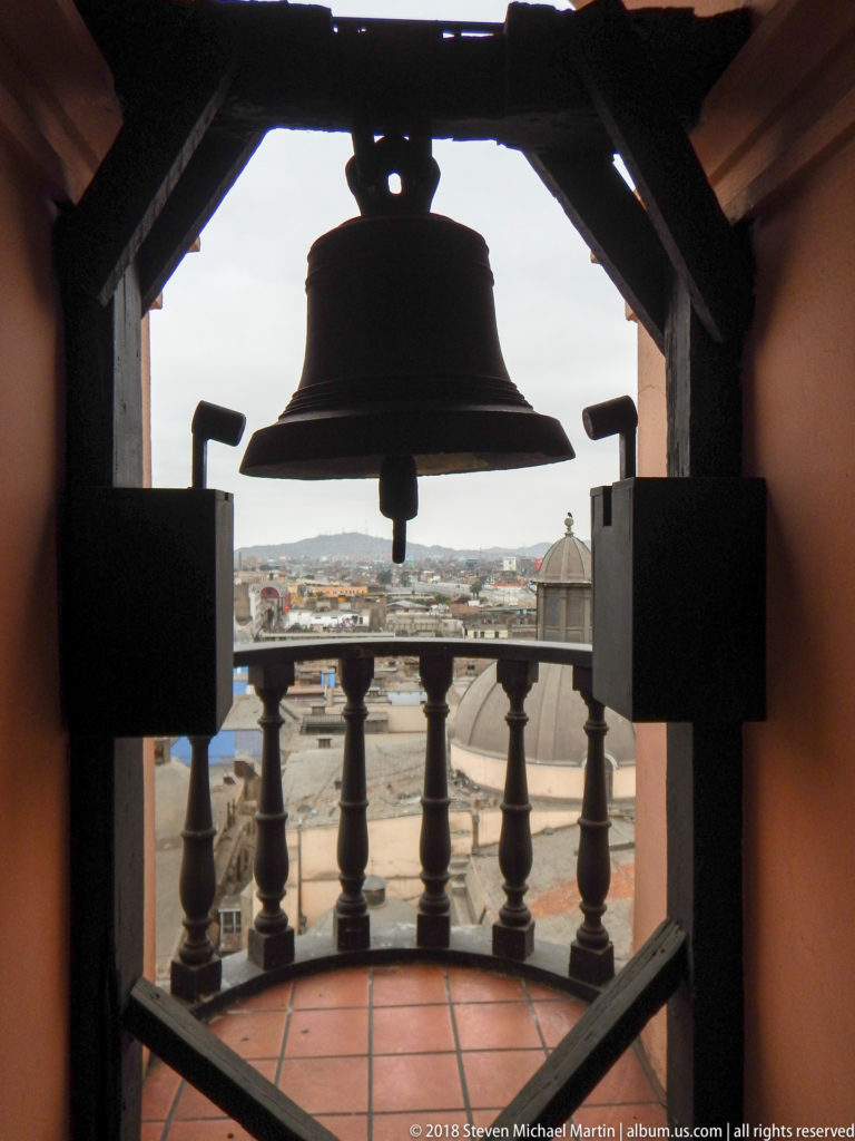 Bell suspended in a heavy wooden frame inside the tower, with tiled floor, balustrade, and Lima skyline beyond.