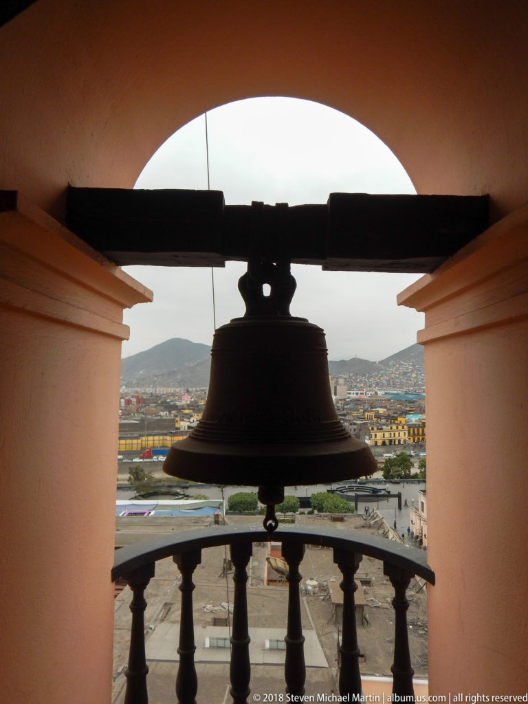 Large bell silhouetted in an arched tower opening, with balustrade, rooftops, and hills of Lima beyond.
