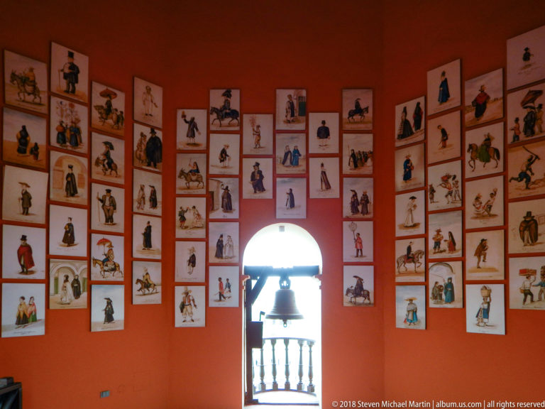 Curved orange tower wall covered with small historical costume prints around an arched opening framing a bell.