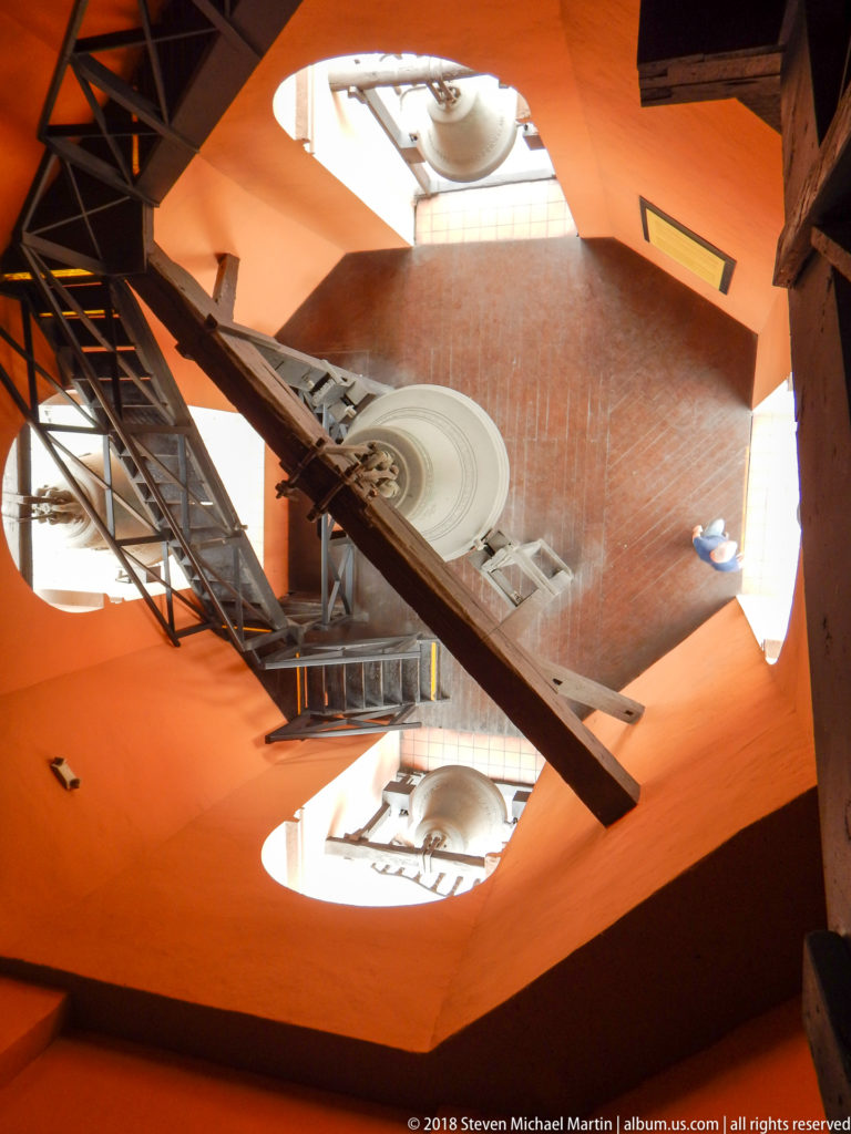 Diagonal overhead view of Santo Domingo bell tower with central bell, surrounding bells, stairs, beams, and orange walls.