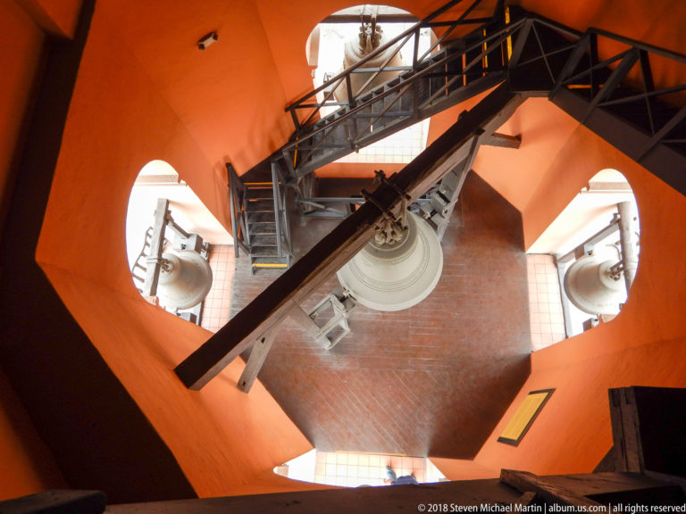 High downward view inside the orange bell tower with several bells, metal stairways, and heavy crossing beams.