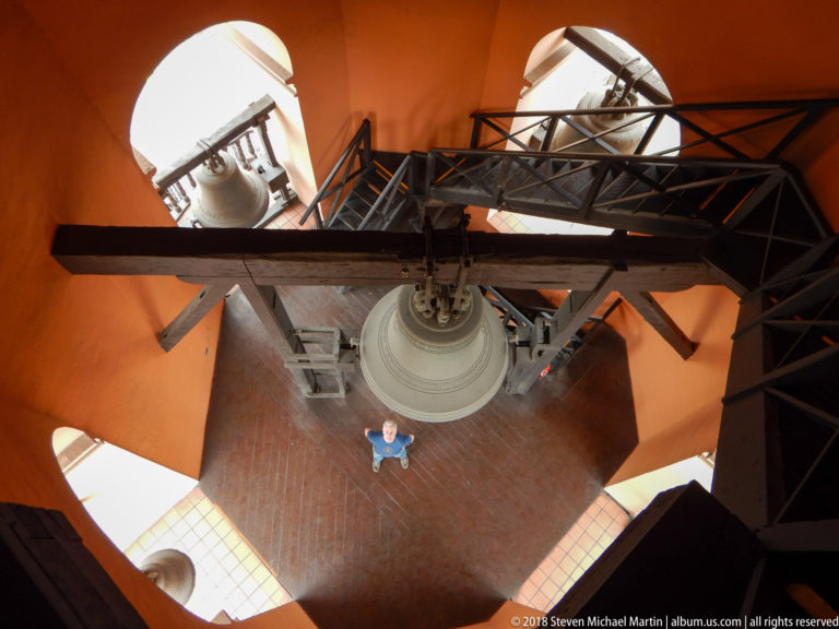 Downward view inside Santo Domingo’s bell tower with crossing beams, metal stairs, large bell, orange walls, and visitor below.