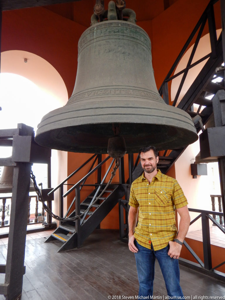 Large bronze church bell suspended in a tower frame above a visitor, with stairs and structural braces behind.