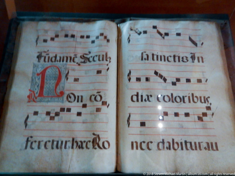 Close-up of choir-book pages with large black musical notation, red staff lines, Latin text, and red-blue decorated initial.